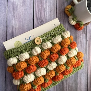 May include: A crochet book sleeve featuring a repeating pattern of small, raised orange and cream-coloured pumpkin shapes on a green background. The sleeve has a single wooden button closure at the top centre and contains a book with visible text.