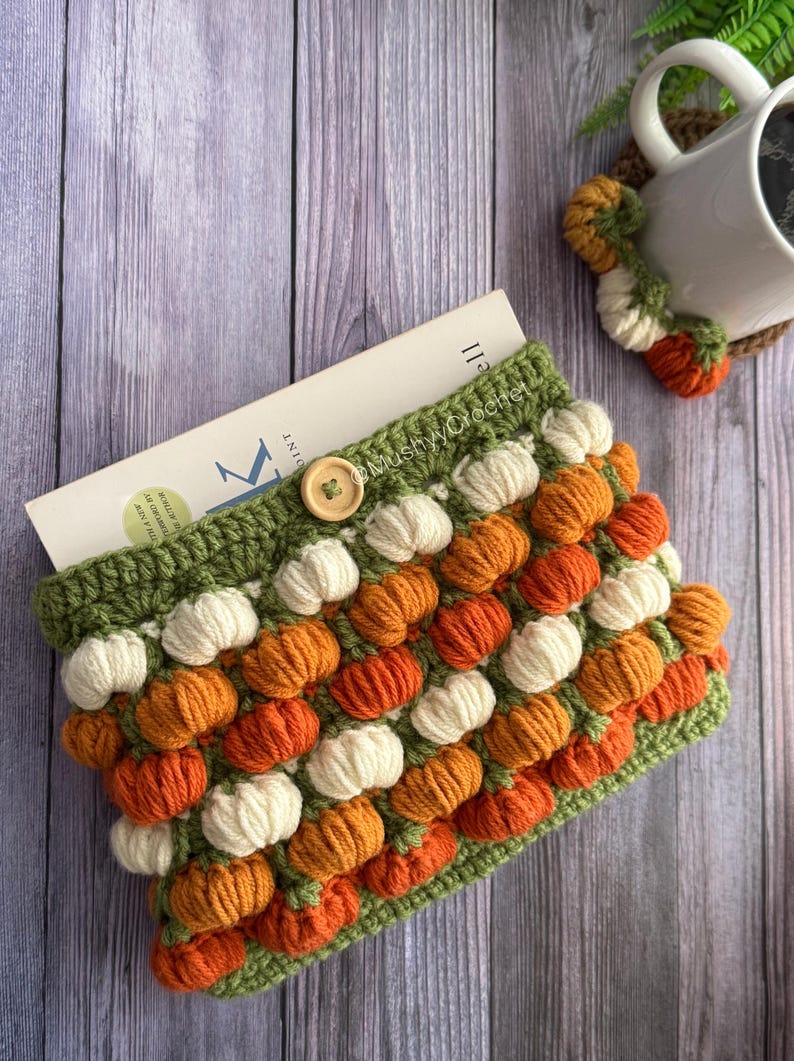 May include: A crochet book sleeve featuring a repeating pattern of small, raised orange and cream-coloured pumpkin shapes on a green background. The sleeve has a single wooden button closure at the top centre and contains a book with visible text.
