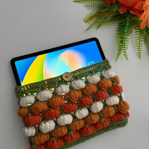 May include: A crochet tablet sleeve featuring a repeating pattern of small white and orange pumpkins on a green background, secured with a single wooden button. The sleeve holds a tablet displaying a colourful abstract screen, with the text @Mushyycrochet.