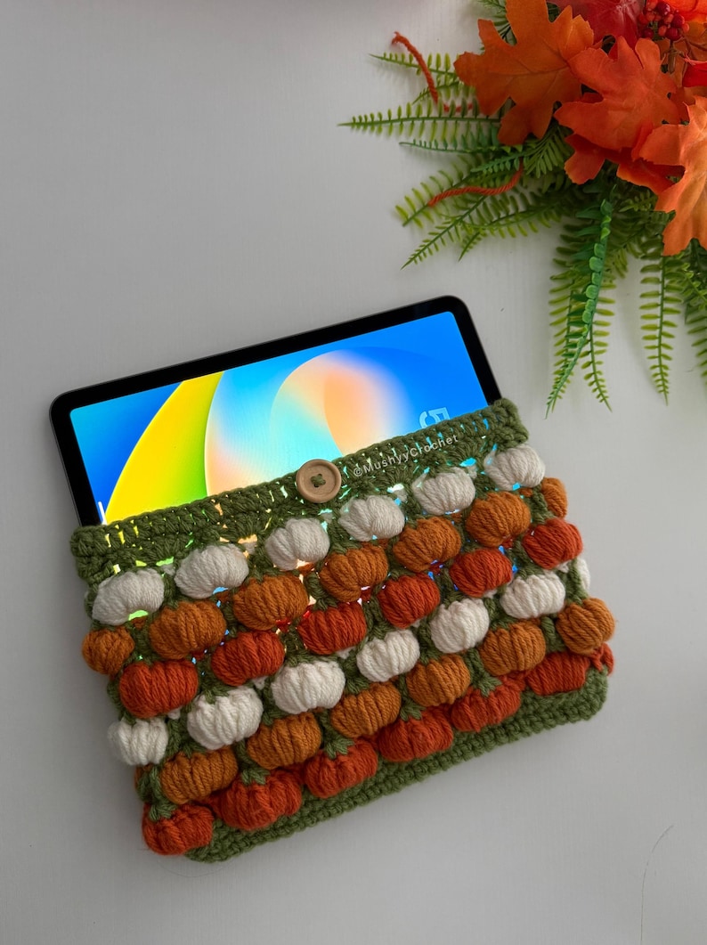 May include: A crochet tablet sleeve featuring a repeating pattern of small white and orange pumpkins on a green background, secured with a single wooden button. The sleeve holds a tablet displaying a colourful abstract screen, with the text @Mushyycrochet.
