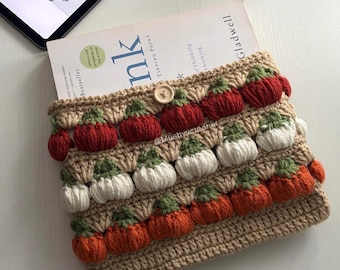 May include: A beige crochet pouch featuring rows of small, textured, three-dimensional pumpkins in red, white, and orange with green stems. The pouch is secured with a single light-coloured wooden button at the top centre and contains a book.