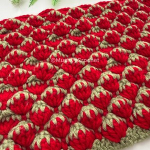 May include: A crocheted textile featuring a repeating pattern of red strawberry-like shapes with light green tops. The fabric has a textured, three-dimensional appearance. The text MushyyCrochet is visible in white lettering across the center of the piece.