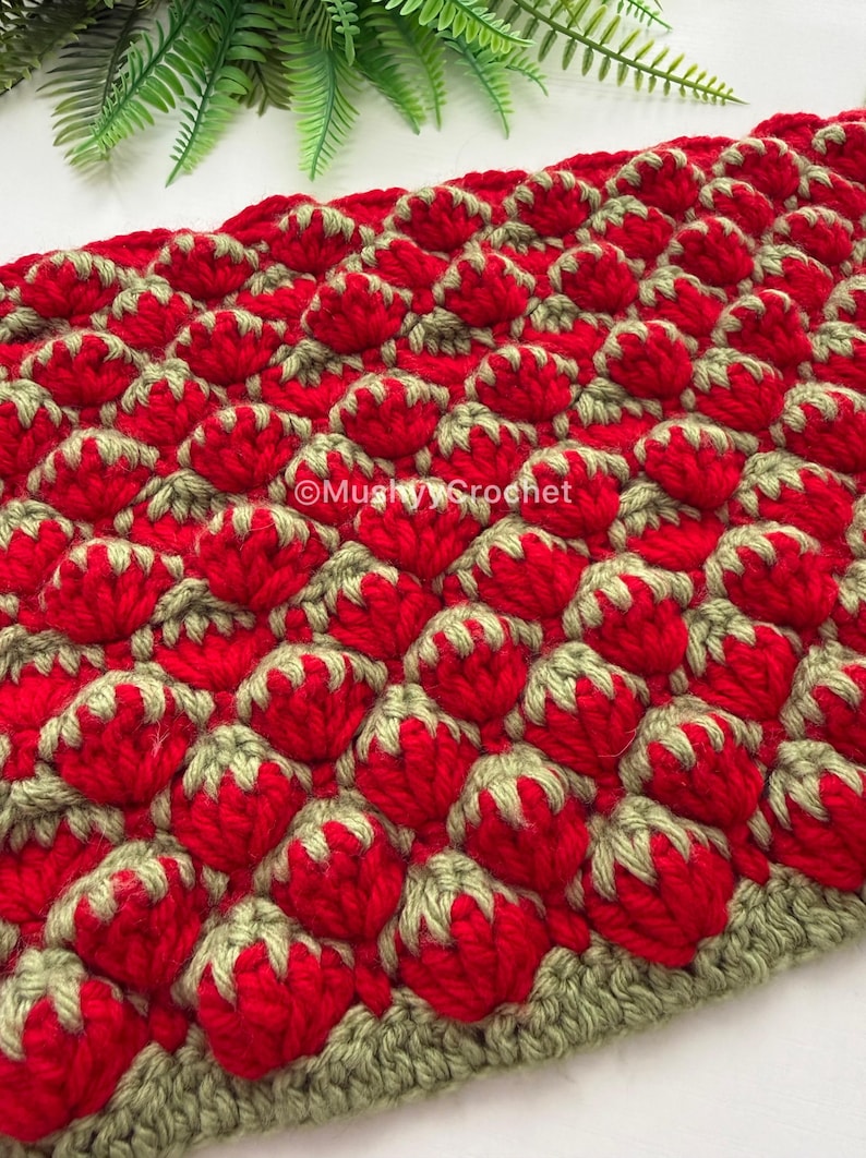 May include: A crocheted textile featuring a repeating pattern of red strawberry-like shapes with light green tops. The fabric has a textured, three-dimensional appearance. The text MushyyCrochet is visible in white lettering across the center of the piece.