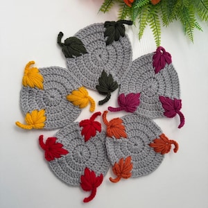 May include: Five circular gray crochet coasters, each featuring three small, textured leaves in different colors: dark green, yellow, magenta, red, and orange. The coasters are arranged in a cluster, showcasing the detailed yarn stitching.