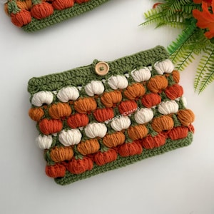 May include: A rectangular crochet pouch featuring a repeating pattern of small, raised white and orange pumpkin-shaped stitches on a green base. The pouch includes a single wooden button closure at the top centre and the text MushyyCrochet.