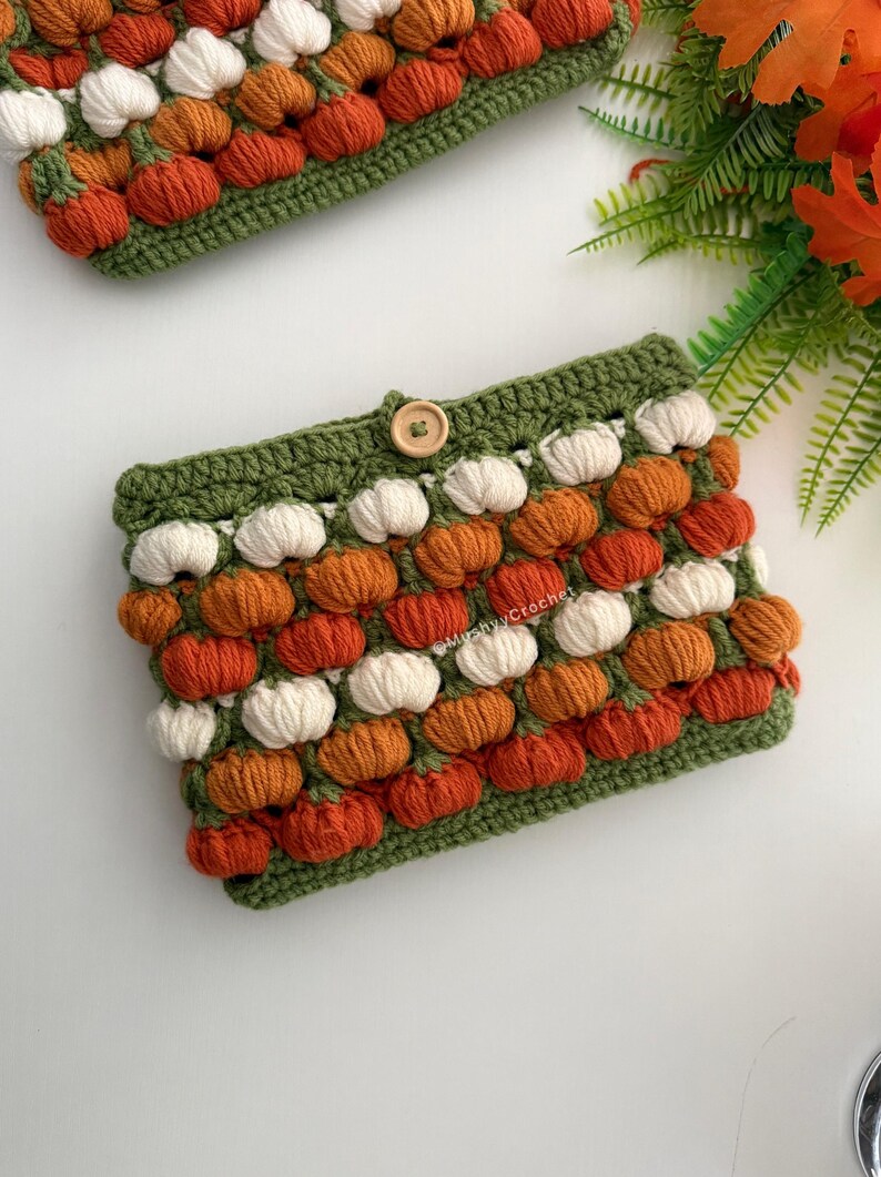 May include: A rectangular crochet pouch featuring a repeating pattern of small, raised white and orange pumpkin-shaped stitches on a green base. The pouch includes a single wooden button closure at the top centre and the text MushyyCrochet.