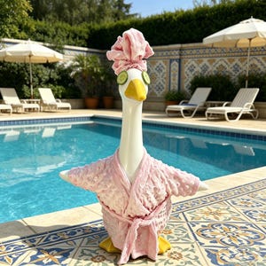 May include: A whimsical white goose figurine with a yellow beak and feet, wearing a pink spa robe, head wrap, and cucumber slices over its eyes. The goose is posed by a swimming pool, suggesting a relaxing spa theme.