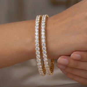 Peut inclure: Deux bracelets rigides dorés ornés d'une rangée continue de pierres rondes, transparentes et facettées sur la surface extérieure. Les bracelets présentent une finition métallique polie et une forme circulaire uniforme.