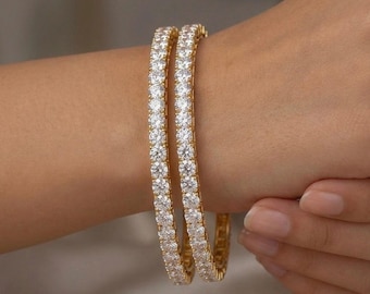 May include: Two gold-toned bangles featuring a continuous row of round, clear, faceted stones set along the outer surface. The bangles have a polished metallic finish and a uniform circular shape, designed to be worn around the wrist.