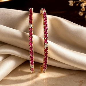 May include: Two gold bangles featuring a continuous row of marquise-cut vibrant pink stones. Each bangle is accented with small, clear, round-cut stones placed at intervals along the band, creating a repeating pattern of pink and clear gems.