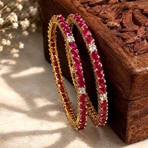 May include: Two gold bangles featuring a continuous row of marquise-cut deep red stones interspersed with small, round, clear white stones. The metal setting is yellow gold, and the design creates a circular, linked pattern of alternating gems.