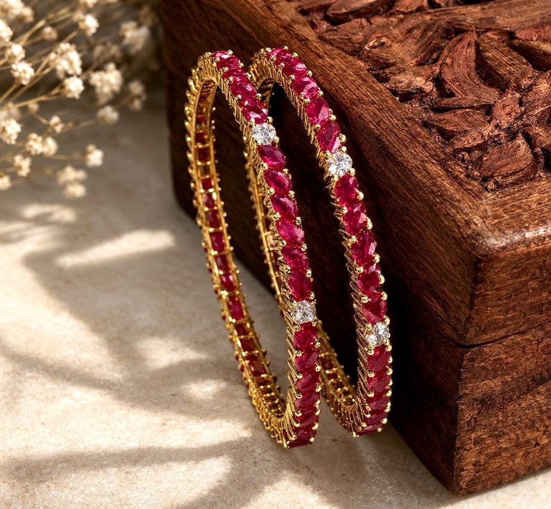 May include: Two gold bangles featuring a continuous row of marquise-cut deep red stones interspersed with small, round, clear white stones. The metal setting is yellow gold, and the design creates a circular, linked pattern of alternating gems.