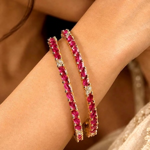 May include: Two gold bangles featuring a continuous row of marquise-cut vibrant pink stones interspersed with small, clear, round-cut white stones. The metal setting is polished gold, creating a bright contrast against the deep pink gemstones.