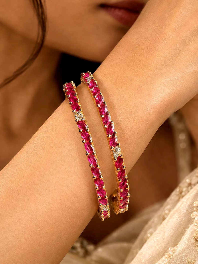 May include: Two gold bangles featuring a continuous row of marquise-cut vibrant pink stones interspersed with small, clear, round-cut white stones. The metal setting is polished gold, creating a bright contrast against the deep pink gemstones.
