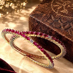 May include: Two gold-toned bangles featuring a continuous row of marquise-cut deep pink stones interspersed with small, round, clear white stones. The metal bands have a textured, ridged edge design that frames the vibrant gemstone inlay.