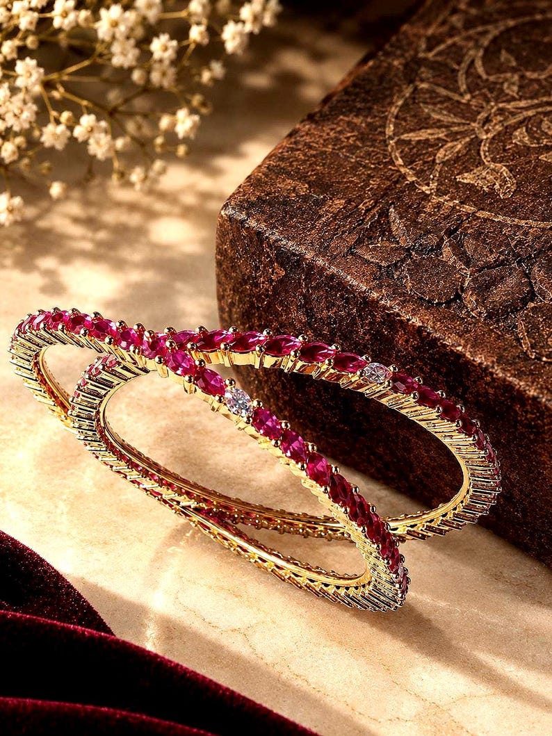 May include: Two gold-toned bangles featuring a continuous row of marquise-cut deep pink stones interspersed with small, round, clear white stones. The metal bands have a textured, ridged edge design that frames the vibrant gemstone inlay.