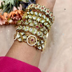 Peut inclure: Plusieurs bracelets dorés ornés de pierres précieuses claires et rouges sont empilés sur un poignet. Les bracelets présentent des motifs détaillés, notamment des motifs floraux et en forme de goutte. Les bijoux sont posés sur un fond doux et neutre.