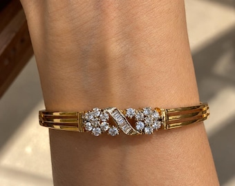 May include: A gold bangle bracelet featuring a central decorative element composed of two clusters of round brilliant-cut diamonds flanking a diagonal row of baguette-cut diamonds. The band consists of three parallel gold wires on each side.