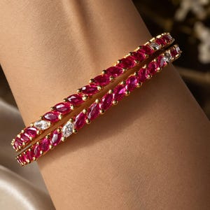 May include: Two gold bangles featuring a continuous row of marquise-cut deep red stones interspersed with occasional clear white stones. The bracelets have a polished gold metal setting that holds each stone securely in a prong-style arrangement.