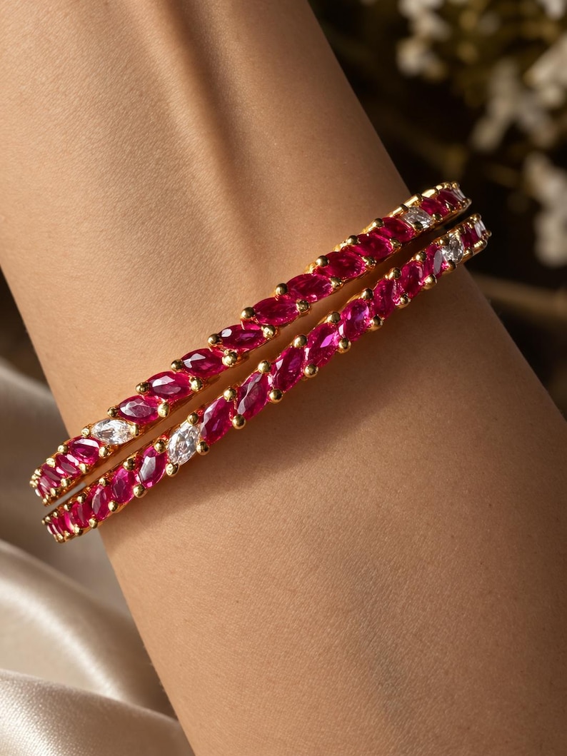 May include: Two gold bangles featuring a continuous row of marquise-cut deep red stones interspersed with occasional clear white stones. The bracelets have a polished gold metal setting that holds each stone securely in a prong-style arrangement.