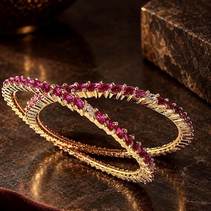 May include: Two gold bangles featuring a continuous row of marquise-cut vibrant pink stones. Each bangle is crafted with a textured gold metal band that holds the stones in a secure setting, creating a circular shape with a polished finish.