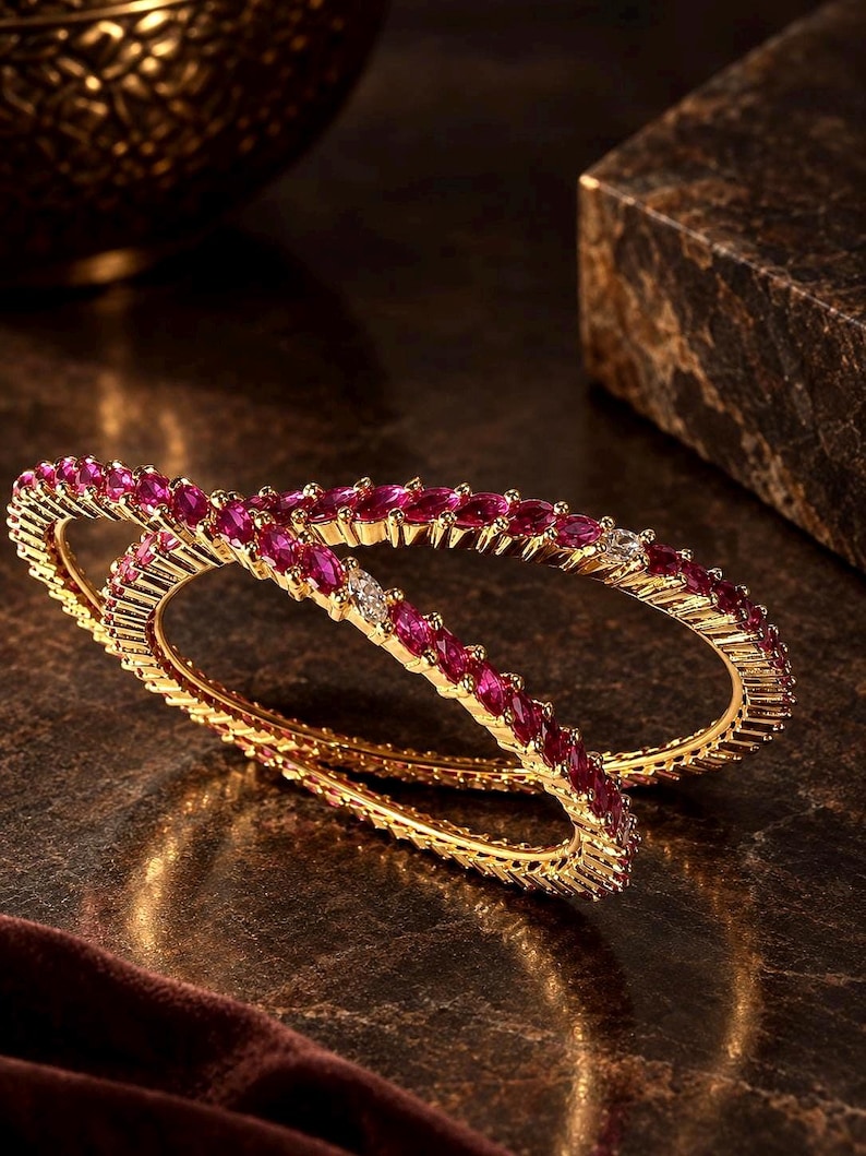 May include: Two gold bangles featuring a continuous row of marquise-cut vibrant pink stones. Each bangle is crafted with a textured gold metal band that holds the stones in a secure setting, creating a circular shape with a polished finish.