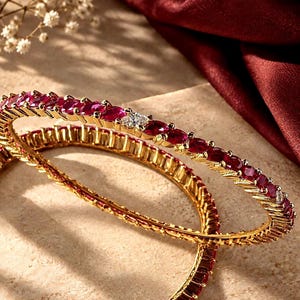 May include: Two gold bangles featuring a continuous row of deep red oval-cut gemstones and occasional clear square-cut stones. The metal bands have a textured, ridged edge design that frames the vibrant red stones along the entire circumference.