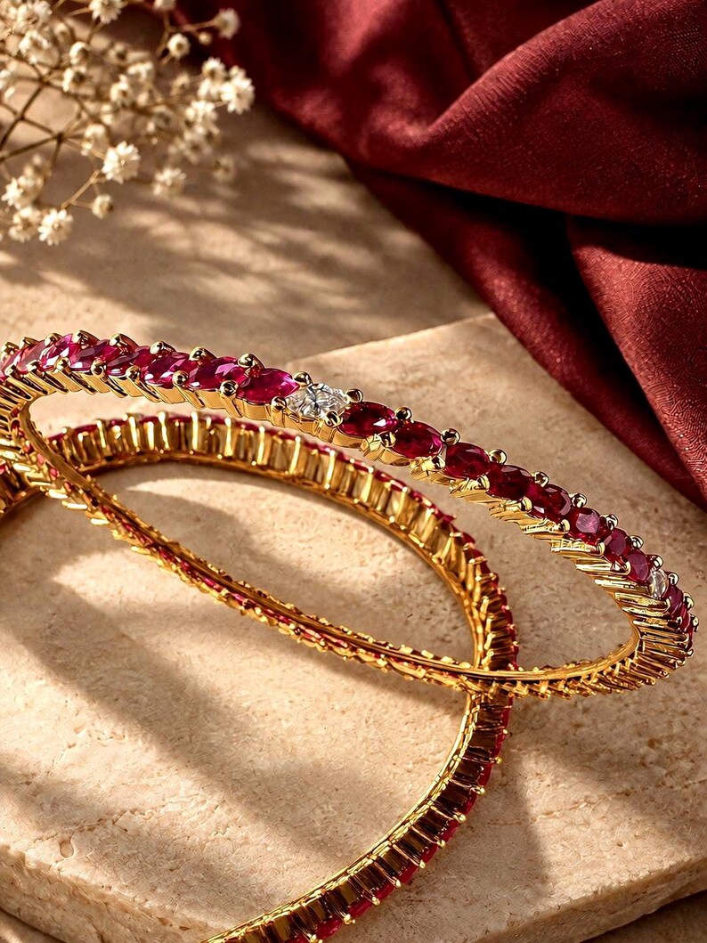 May include: Two gold bangles featuring a continuous row of deep red oval-cut gemstones and occasional clear square-cut stones. The metal bands have a textured, ridged edge design that frames the vibrant red stones along the entire circumference.
