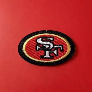 Peut inclure: Un écusson brodé de forme ovale présentant les lettres SF en blanc avec un contour noir. Les lettres sont centrées sur un fond rouge, entourées d'une fine bordure dorée et d'une épaisse bordure noire extérieure avec une finition cousue.
