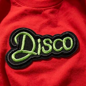 May include: A bright red sweatshirt featuring a black embroidered patch with the word Disco stitched in a vibrant lime green cursive font. The patch has a thick black border and a textured finish, contrasting against the solid red fabric background.