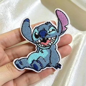 May include: An embroidered patch featuring the character Stitch with blue fur, a light blue chest patch, and a wide, grinning mouth showing white teeth. The character has large ears, one with a pink inner section, and is depicted in a seated pose.