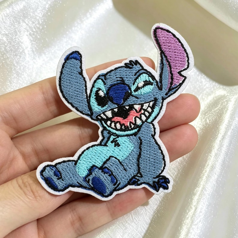 May include: An embroidered patch featuring the character Stitch with blue fur, a light blue chest patch, and a wide, grinning mouth showing white teeth. The character has large ears, one with a pink inner section, and is depicted in a seated pose.