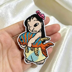 Op de afbeelding: Een geborduurde patch met een stripfiguur met zwart haar en een roze bloem, die een grote oranje en blauw gestreepte vis vasthoudt. Het figuur draagt een lichtblauwe en gele rok met patroon, met witte omlijningen van de vorm.