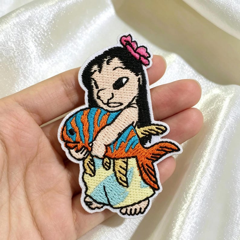 Op de afbeelding: Een geborduurde patch met een stripfiguur met zwart haar en een roze bloem, die een grote oranje en blauw gestreepte vis vasthoudt. Het figuur draagt een lichtblauwe en gele rok met patroon, met witte omlijningen van de vorm.
