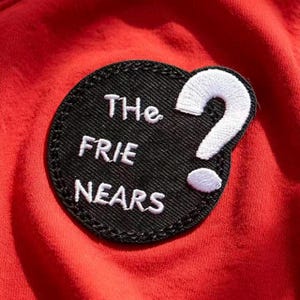 May include: A circular black embroidered patch attached to a red fabric background. The patch features white stitched text that reads "THe FRIE NEARS" and a large white embroidered question mark positioned to the right of the text.