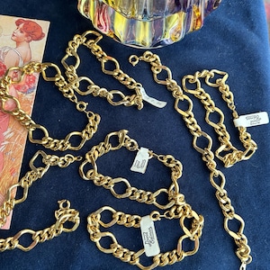 May include: A collection of gold-toned metal chain bracelets featuring alternating oval and curb links. Several bracelets include small white rectangular tags with the text David Salmon printed in black. The jewellery is arranged on a dark blue surface.
