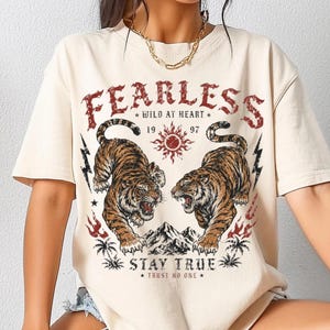 Puede incluir: Una camiseta de manga corta de color crema con un estampado gráfico de dos tigres frente a frente sobre una cordillera. El texto dice FEARLESS, WILD AT HEART, 1997, STAY TRUE y TRUST NO ONE, acompañado de iconos de sol, rayos y palmeras.