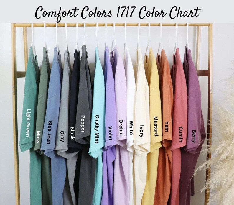 May include: A color chart of Comfort Colors 1717 t-shirts, showcasing a range of hues from light green to berry. Each shirt hangs on a white hanger, with color names like "Mustard" and "Orchid" displayed below.
