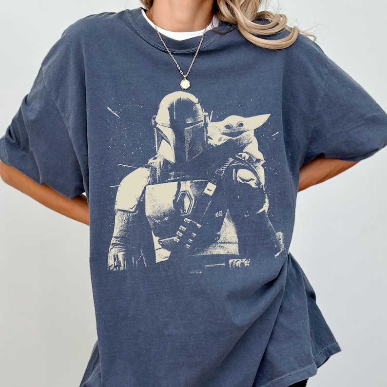 May include: A navy blue t-shirt featuring a graphic of The Mandalorian and Grogu. The design is in a vintage style, with the characters in a light beige color. The shirt has short sleeves and a crew neck.