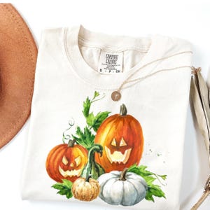 May include: A cream-colored t-shirt with a Comfort Colors label at the neck, featuring a watercolor-style graphic of three carved jack-o'-lanterns and one white pumpkin surrounded by green leaves and vines on the front of the garment.