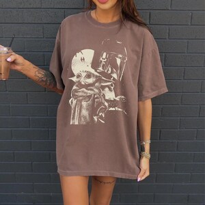 May include: Brown t-shirt featuring a graphic of Grogu and the Mandalorian. The design is in a cream color, set against a brown background. The shirt has short sleeves and a relaxed fit.