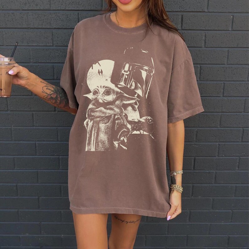 May include: Brown t-shirt featuring a graphic of Grogu and the Mandalorian. The design is in a cream color, set against a brown background. The shirt has short sleeves and a relaxed fit.