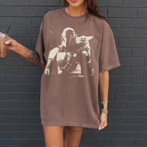 May include: Brown t-shirt featuring a graphic of a Mandalorian and Grogu. The design is in a light brown color, contrasting with the darker brown of the shirt. The shirt has short sleeves and a relaxed fit, suitable for casual wear.