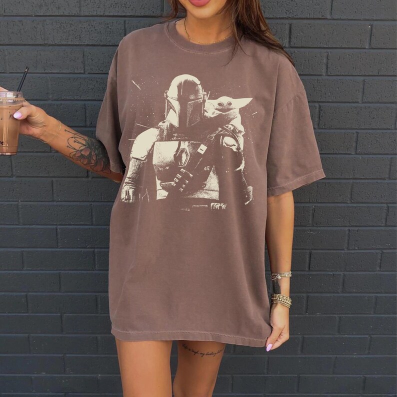 May include: Brown t-shirt featuring a graphic of a Mandalorian and Grogu. The design is in a light brown color, contrasting with the darker brown of the shirt. The shirt has short sleeves and a relaxed fit, suitable for casual wear.