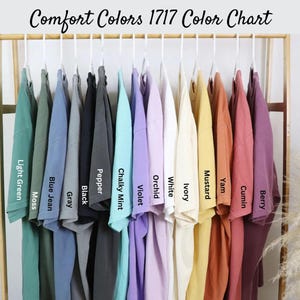 May include: A color chart of Comfort Colors 1717 t-shirts, showcasing a range of hues from light green to berry. Each shirt hangs on a white hanger, with color names like "Mustard" and "Orchid" displayed below.