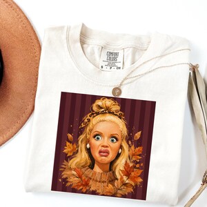 Könnte beinhalten: Ein cremefarbenes T-Shirt mit einem Grafikdruck einer blonden Person mit überraschtem Gesichtsausdruck, die ein Stirnband mit Leopardenmuster und einen braunen Pullover trägt, umgeben von Herbstblättern vor einem dunkelrot-schwarz gestreiften Hintergrund.