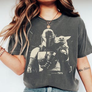 May include: Dark gray t-shirt featuring a black and white graphic of The Mandalorian and Grogu. The design shows the Mandalorian in his armor with Grogu. The shirt has a vintage, worn look.