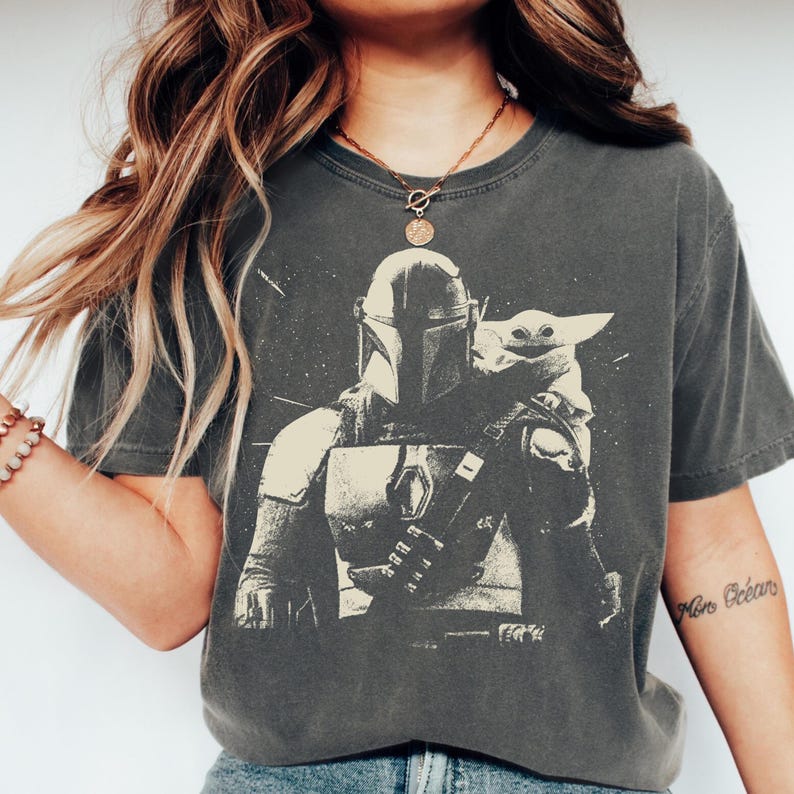 May include: Dark gray t-shirt featuring a black and white graphic of The Mandalorian and Grogu. The design shows the Mandalorian in his armor with Grogu. The shirt has a vintage, worn look.
