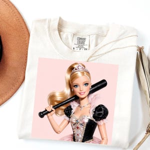 Könnte beinhalten: Ein cremefarbenes Kurzarm-T-Shirt mit einem Comfort Colors-Etikett am Halsausschnitt. Der Aufdruck zeigt eine blonde Puppe mit Tiara und einem schwarz-rosafarbenen Kleid, die einen schwarzen Baseballschläger vor einem hellrosa Hintergrund hält.