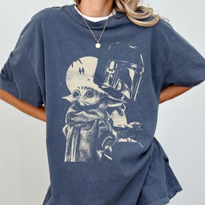 May include: Dark blue t-shirt featuring a graphic of Grogu and the Mandalorian helmet. The design is in a faded, vintage style. The shirt has a crew neck and short sleeves.